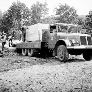 Truck transports lumber on a dirt road, workers present.