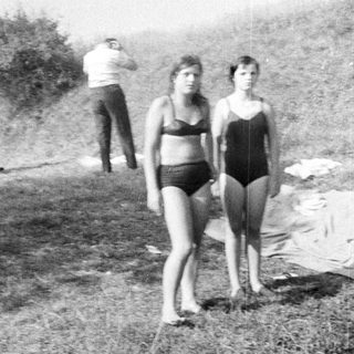 Here's a brief summary of the image: People relaxing near a building, two girls in swimsuits.