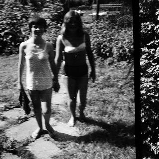 Here are a few brief summaries of the image: Two people walk a path near foliage. Black and white photo shows people