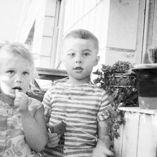 Two young children stand on a porch, looking at the camera.