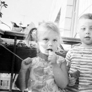 Two young children stand on a porch, looking at the camera.