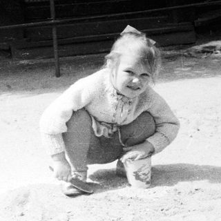 Here's a brief summary of the image: Child kneeling outdoors, in front of a building.