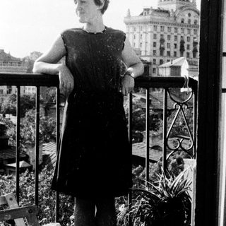 Woman stands on balcony overlooking a building and city.