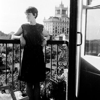 Woman stands on balcony overlooking a building and city.