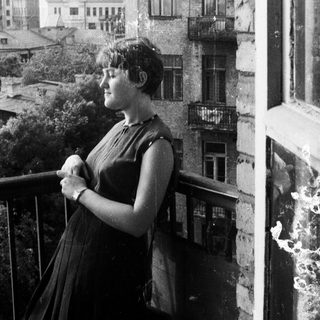 Woman stands on balcony, overlooking a city skyline.