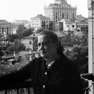Woman stands on balcony overlooking a city and large building.