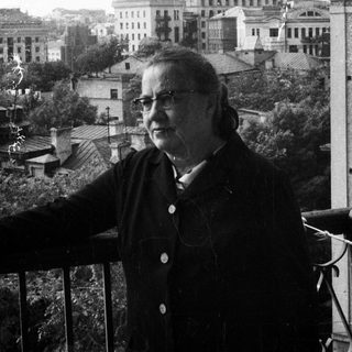 Woman stands on balcony overlooking a city and large building.