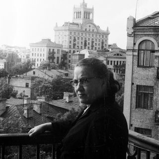 Woman stands on balcony overlooking city with grand building.