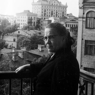 Woman stands on balcony overlooking city with grand building.