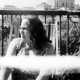 Woman sits outdoors, overlooking a building with a tower.