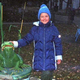 A child stands next to a frog statue in a garden.