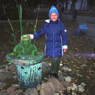 A child stands next to a frog statue in a garden.