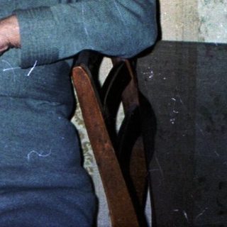Older woman seated in a room with patterned wallpaper.