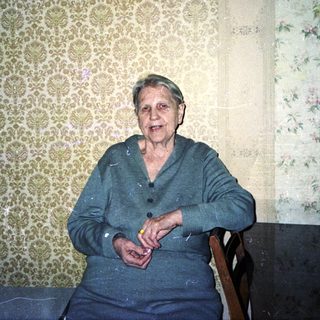 Older woman seated in a room with patterned wallpaper.