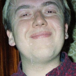 Man in plaid shirt stands in front of patterned wallpaper.