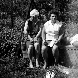 Here's a brief summary of the image: Two women seated on a wooden bench; another reclined nearby.