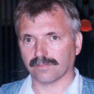 Man with mustache stands in a wooden-paneled room.