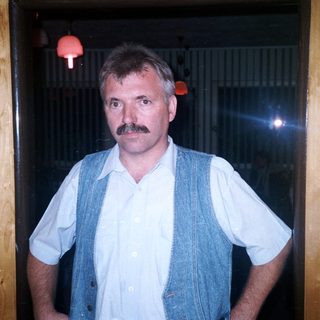 Man with mustache stands in a wooden-paneled room.