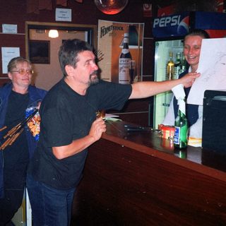 Here's a brief summary of the image: Men and a woman standing at a bar, looking at a paper.
