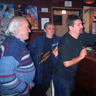 Here's a brief summary of the image: Men and a woman standing at a bar, looking at a paper.