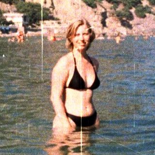 Woman stands in water, mountain backdrop, beach in background.