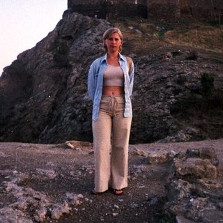 Woman stands before a castle on a rocky hillside.