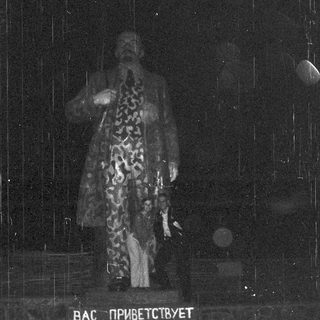 Man in patterned coat stands before Cyrillic text on a stage.