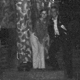 Man in patterned coat stands before Cyrillic text on a stage.
