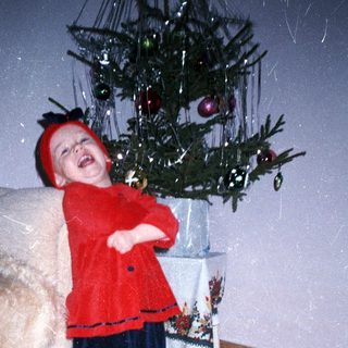 Child in festive clothing stands near a decorated Christmas tree.