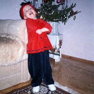 Child in festive clothing stands near a decorated Christmas tree.