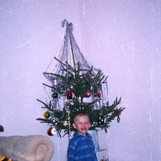 Child stands beneath a small, decorated Christmas tree.