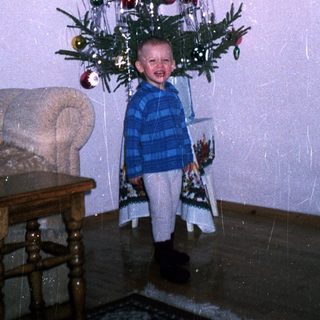 Child stands beneath a small, decorated Christmas tree.