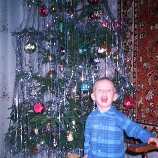 Child stands near a Christmas tree with tinsel.
