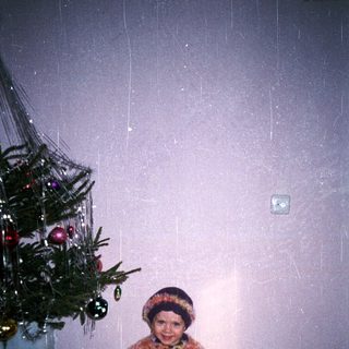 Child stands near a Christmas tree, wearing a furry sweater.