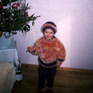 Child stands near a Christmas tree, wearing a furry sweater.
