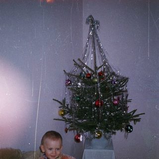 Child sits near a tinsel Christmas tree in a room.