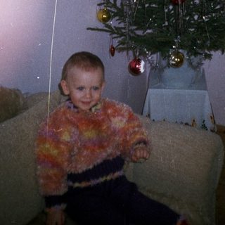 Child sits near a tinsel Christmas tree in a room.