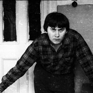 Man in plaid shirt stands near a door, looking forward.