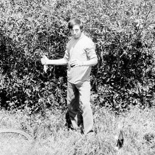 Man stands in foliage, holding something, near a bicycle.