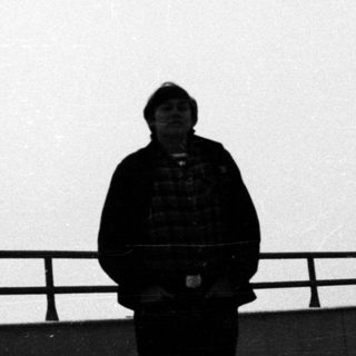 Person stands near a railing against a plain background.