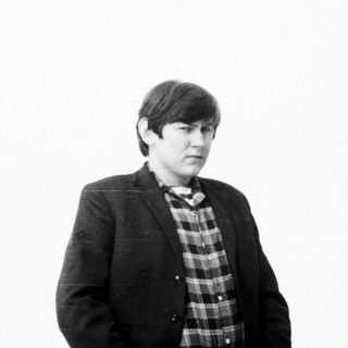 Here's a brief summary of the image: Man in a blazer and plaid shirt stands looking away.