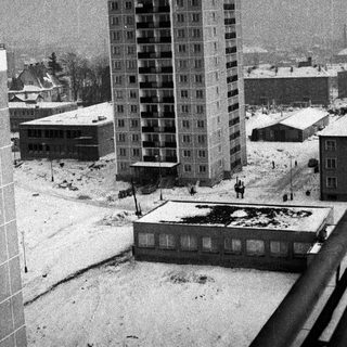 Snowy cityscape featuring a tall apartment building and other structures.