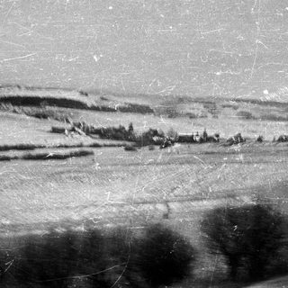 Hilly landscape with trees, visible damage/scratches on the photograph.