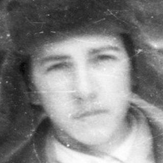 Man in a fur hat stands before a wooden building.