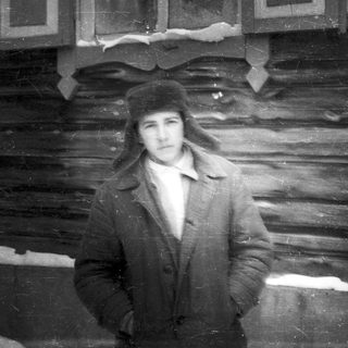 Man in a fur hat stands before a wooden building.