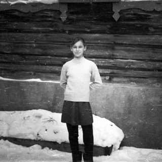 Here's a brief summary of the image: Young girl stands in snow, next to a wooden structure.
