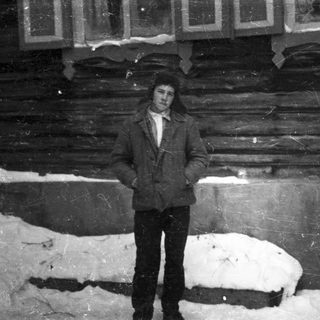 Man stands in snow, positioned in front of a log building.