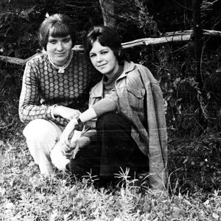 Two young women sit near a tree in a field.