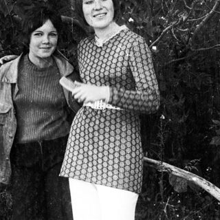 Two women stand outdoors, dressed in vintage clothing.