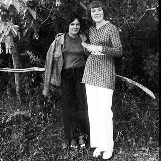 Two women stand outdoors, dressed in vintage clothing.